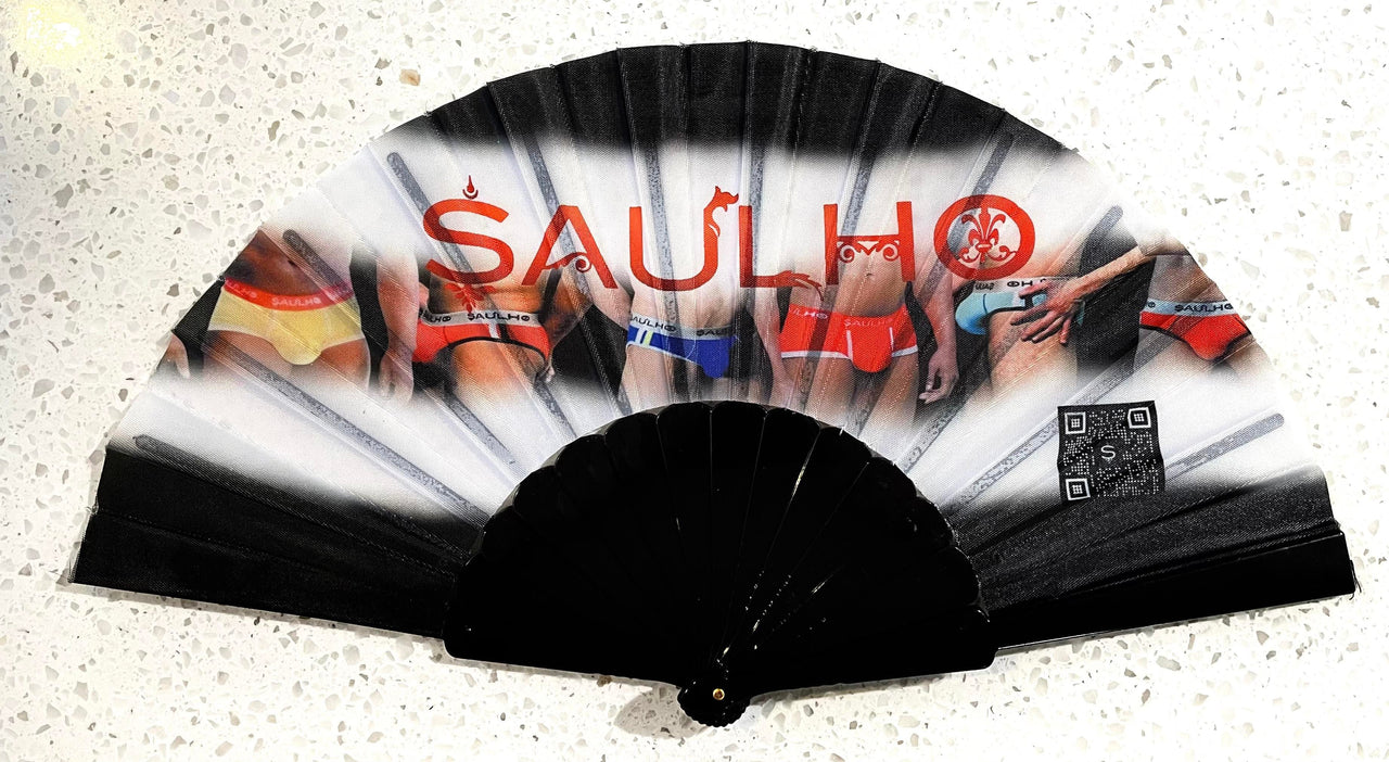 A retractable hand fan that has a black plastic base, connected by a brass colored aluminum screw.  The body of the fan is primarily black in color, however it has white clouds going all the way across .  in the middle of those clouds appears the midsection of 6 men, who are each wearing a different color and model of SAULHO underwear.